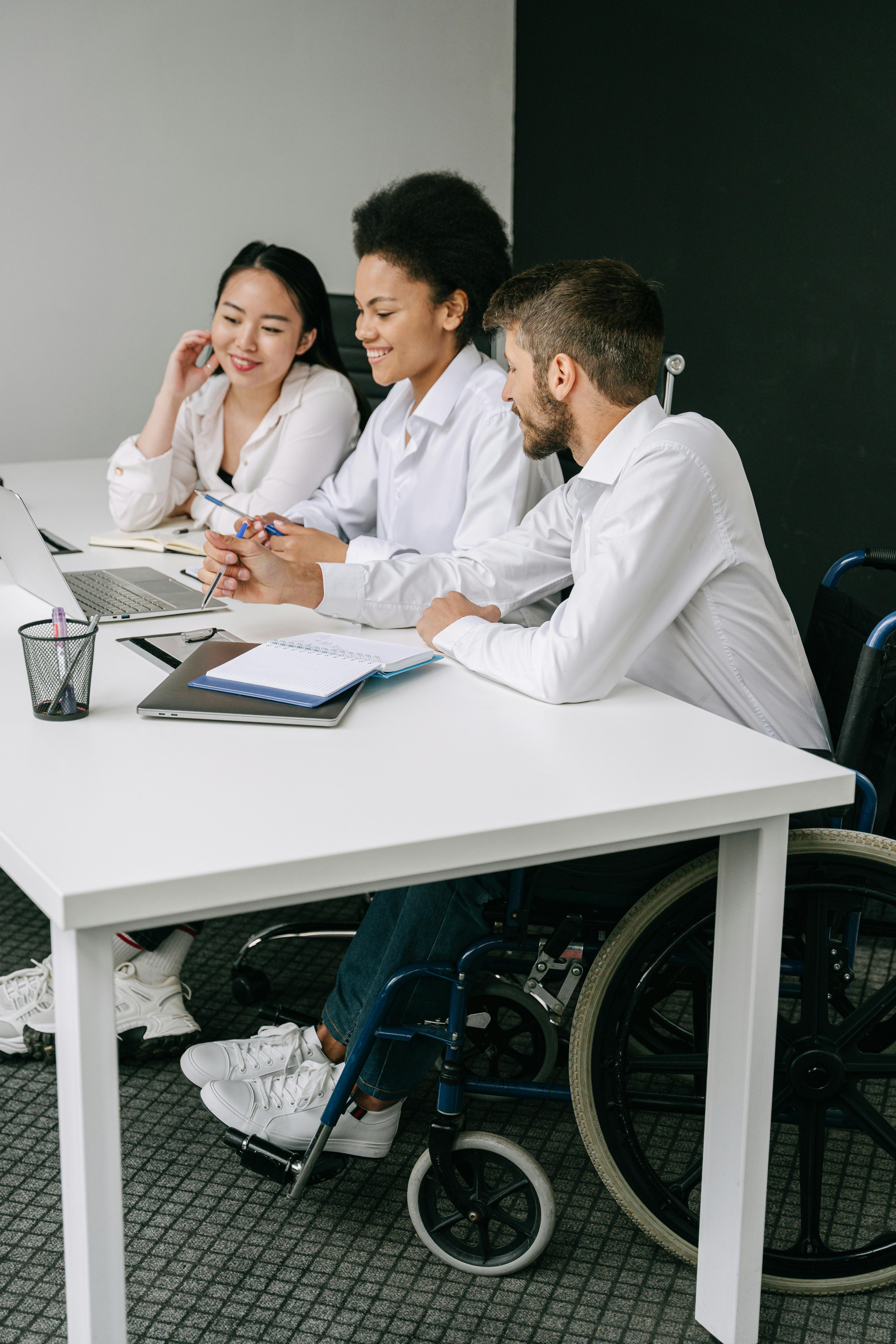 Three colleagues in white shirts collaborate at a desk with laptops and notebooks; one team member uses a wheelchair.